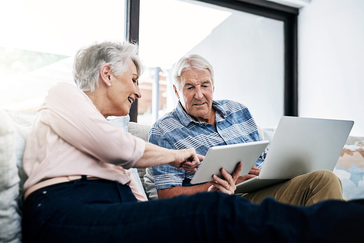 Senior man and woman sit on a couch and review information on a laptop and tablet screen together