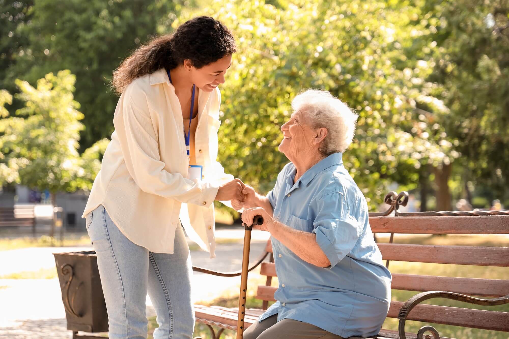 Senior woman sits on an outdoor bench smiling up at younger female caretaker